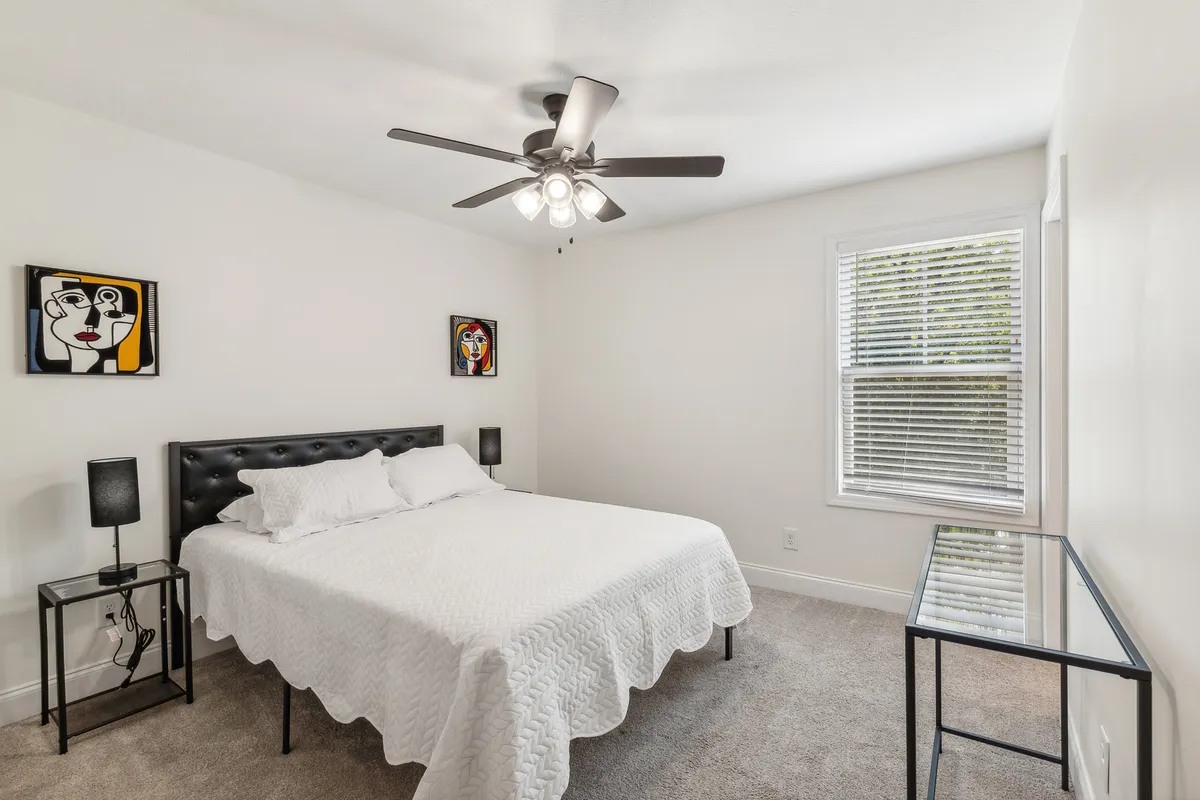 A serene bedroom featuring a comfortable bed with crisp white bedding, abstract wall art above the headboard, black bedside tables, and a window with blinds that filters in soft natural light. The room exudes a calm, inviting atmosphere ideal for relaxation.