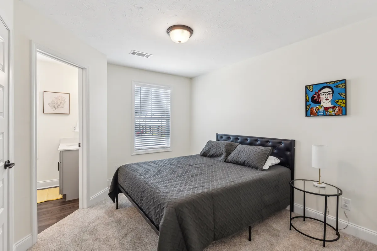 A cozy bedroom showcasing a bed with a dark quilt, an eye-catching art piece on the wall, and a window with blinds. The room's design is intimate and inviting, perfect for relaxation, reading, or quiet reflection.
