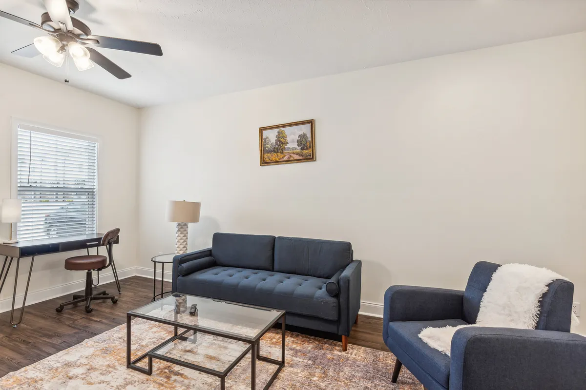 The image showcases a bright, inviting living space featuring a plush sofa, a modern coffee table perfect for casual gatherings, a functional desk and chair ideal for remote work or study, and an eye-catching painting that adds a touch of artistic flair. The arrangement suggests a space equally suited for relaxation, productivity, and socializing.