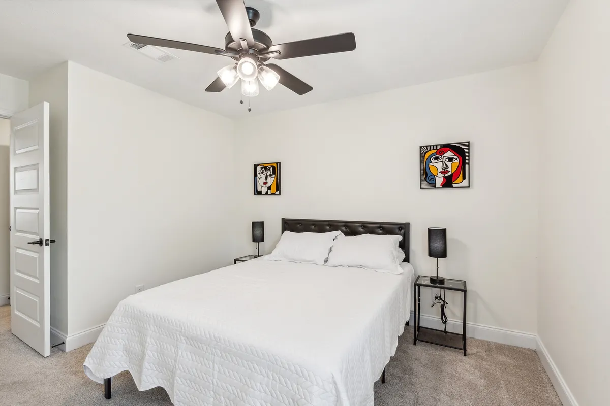 This image displays a comforting bedroom with a bed dressed in crisp white bedding, inviting artwork on the walls, a ceiling fan for added comfort, and nightstands with softly glowing lamps. The room is well-lit with natural light, conveying a serene and homely atmosphere.