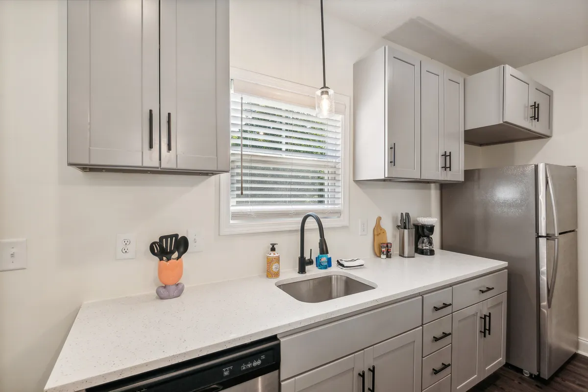 This image highlights the kitchen sink, spacious countertop, modern appliances, and a window with blinds that lets in natural light. The setup feels inviting for both cooking and socializing, with a glimpse of the connected living space, reinforcing the kitchen’s central role in the home.
