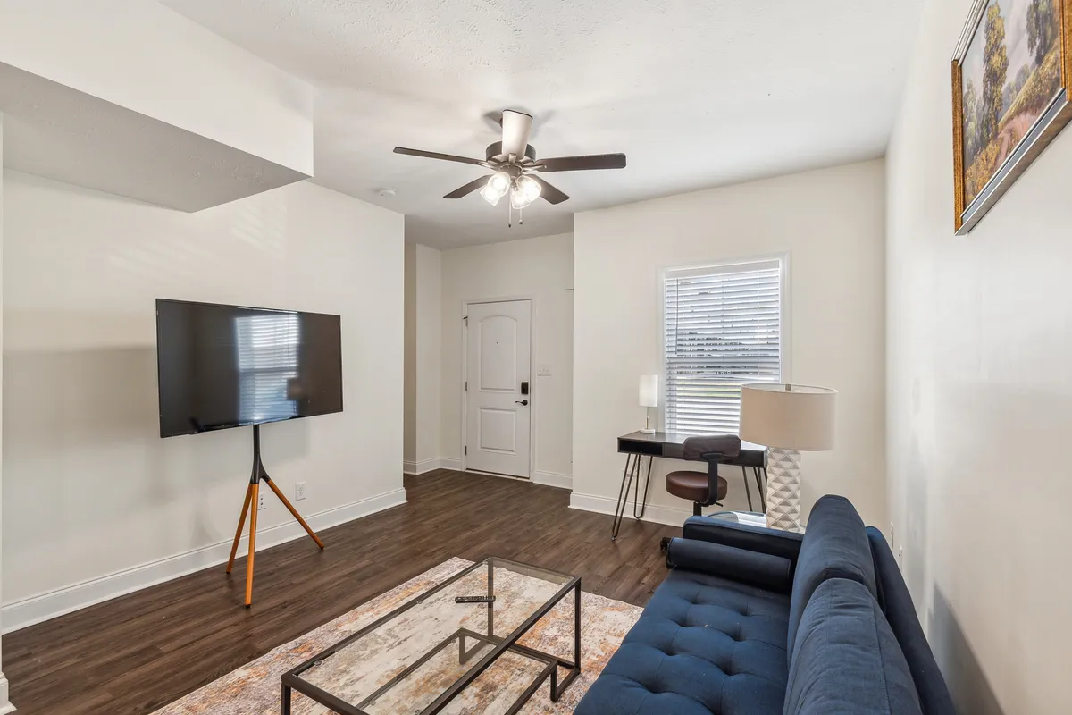 A bright, welcoming view from the front door, featuring a comfortable sofa, coffee table, TV, and lamp, all set in a cozy living area that feels immediately inviting. The front door in frame subtly references the entryway.